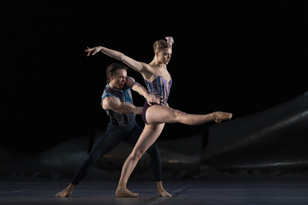 Matthew Ball and Gina Storm-Jensen perform a contemporary pas de deux on a darkly lit stage. Ball lunges and holds Storm-Gensen's hips as she leans forward, her right leg extended devant and her arms lifted up and behind her.