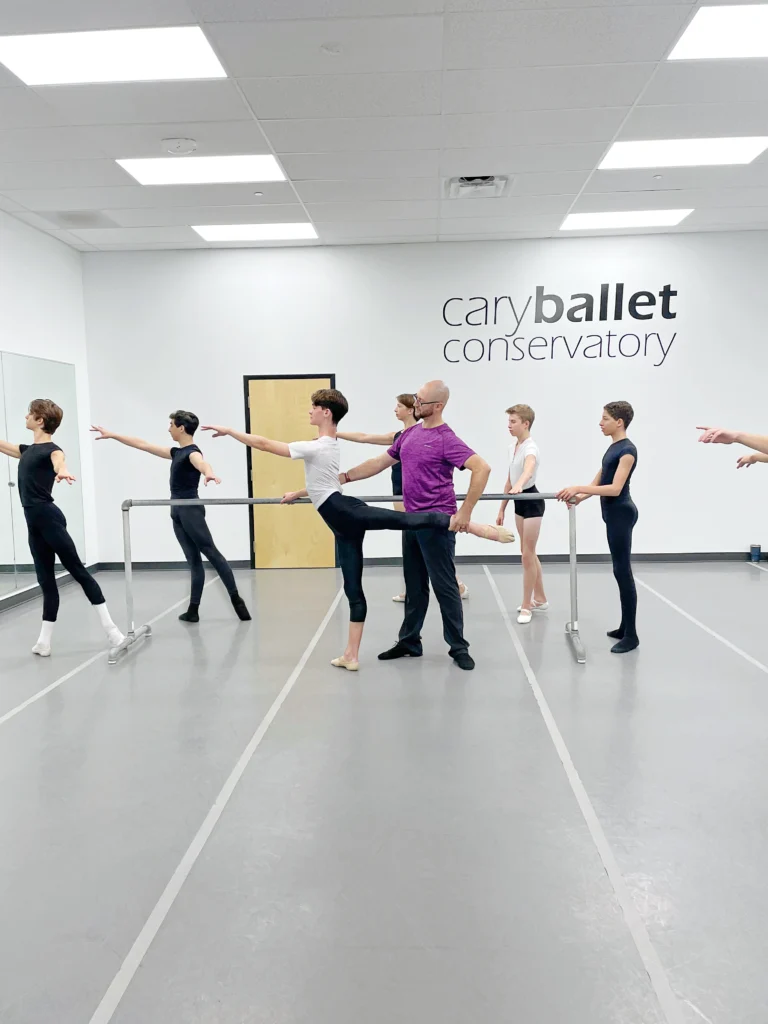 Matthew Donnell, wearing a purple T-shirt and lack pants, adjusts the arabesque of a teenage boy at the barre during a ballet class. Donnell holds the boys left leg up as he poses and corrects his back alignment. Five other teenage male dancers observe, and some practice their arm positions. They male students wear t-shirts, black tights and ballet slippers.