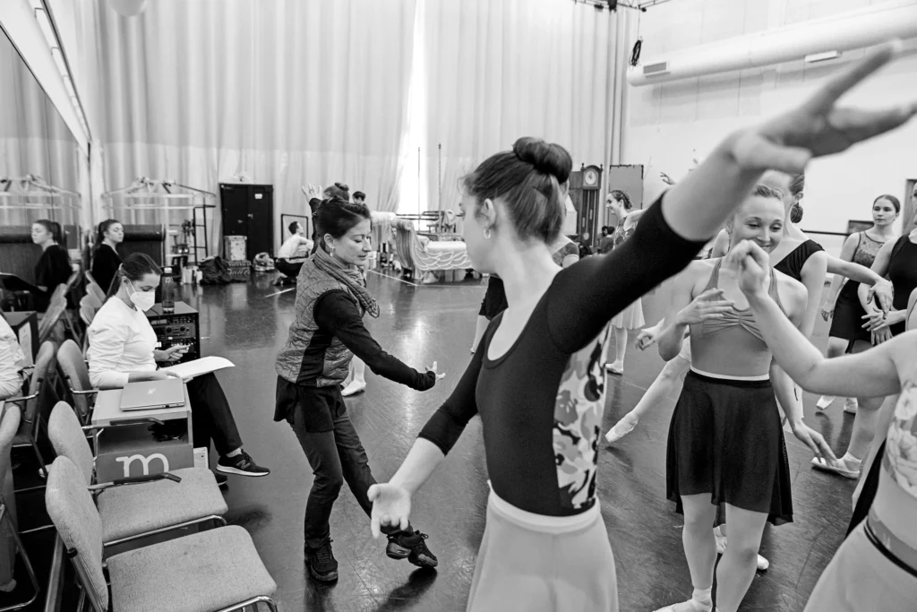 In a large, crowded dance studio, Mikelle Bruzina demonstrates a tendu devant plié with her arms in third position. A group of female dancers watch and copy her, or talk to each other. Bruzina wears puffy vest, dark long-sleeved shirt, dark athletic pants and dark sneakers, while the other women are in leotards, tights, skirts and pointe shoes.