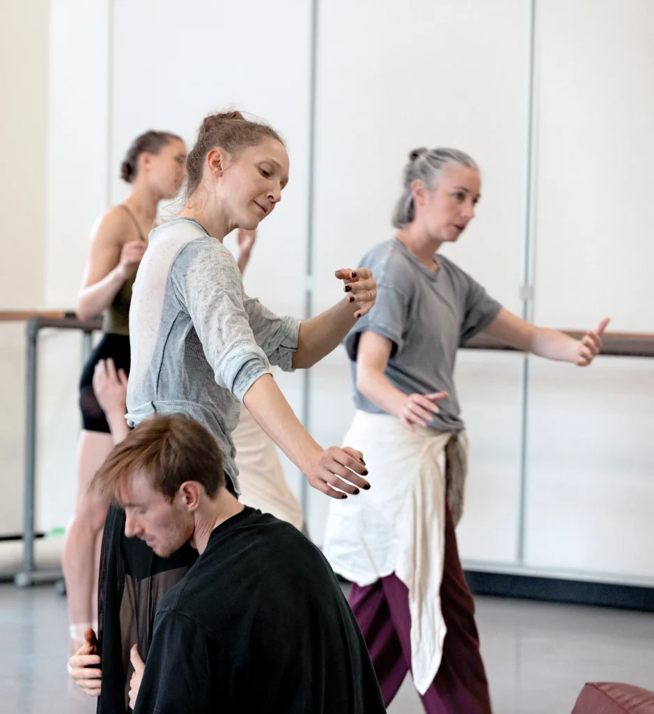 In a large rehearsal studio, Dani Rowe teaches choreography to a male and female duo. The ballerina follows Rowe's movements, bending forward slightly and curving her arms in; her partner kneels down and holds her legs.