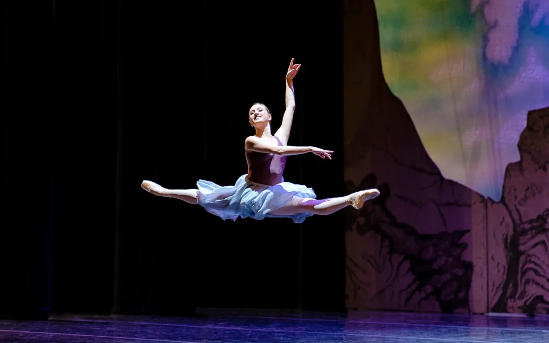 Sophie Williams, wearing a purple leotard and long, lavender skirt, does a giant saut de chat out of the wings on stage right during a performance. She holds her arms in third arabesque and looks out towards the audience with a large smile.