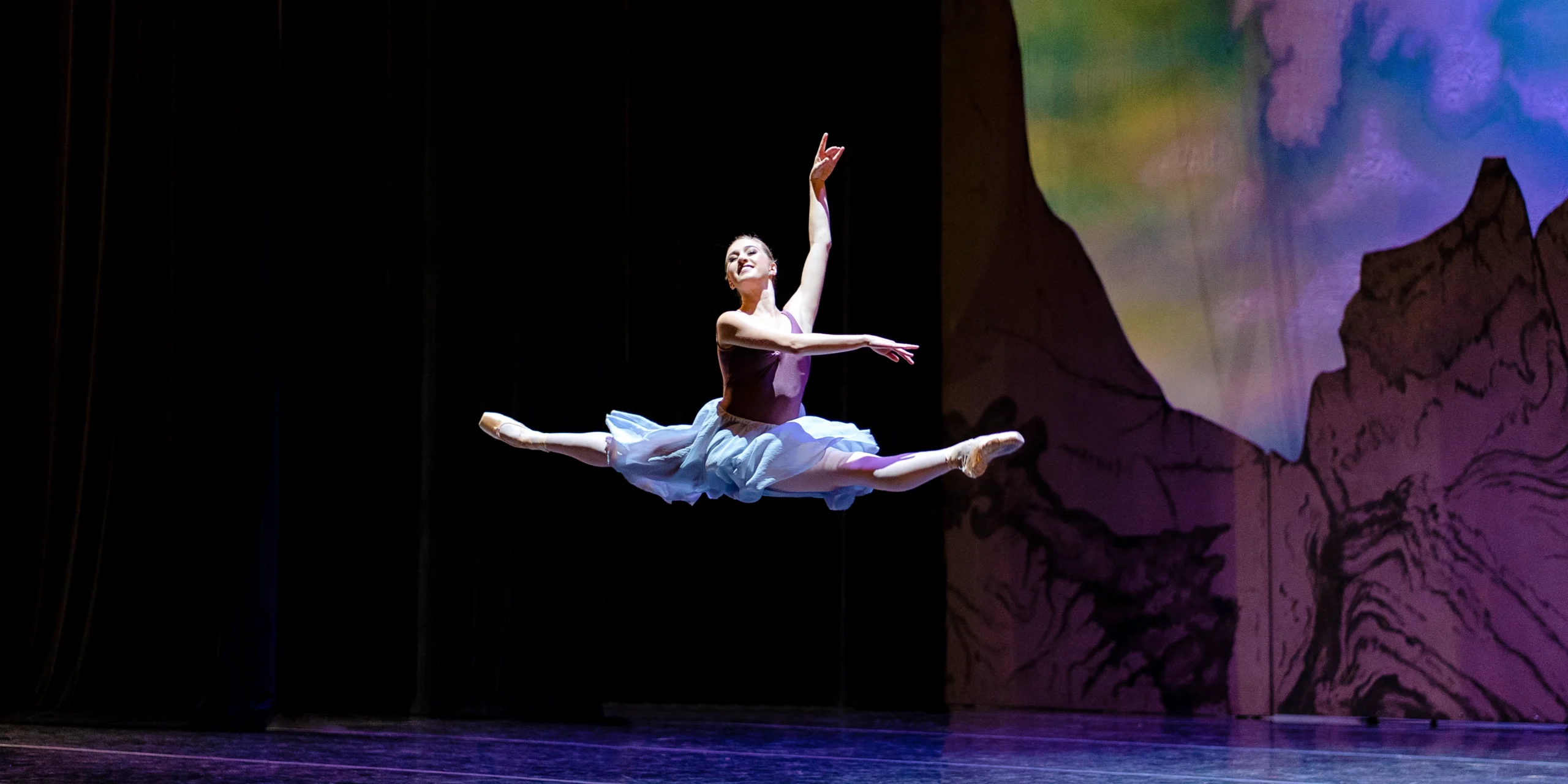Sophie Williams, wearing a purple leotard and long, lavender skirt, does a giant saut de chat out of the wings on stage right during a performance. She holds her arms in third arabesque and looks out towards the audience with a large smile.