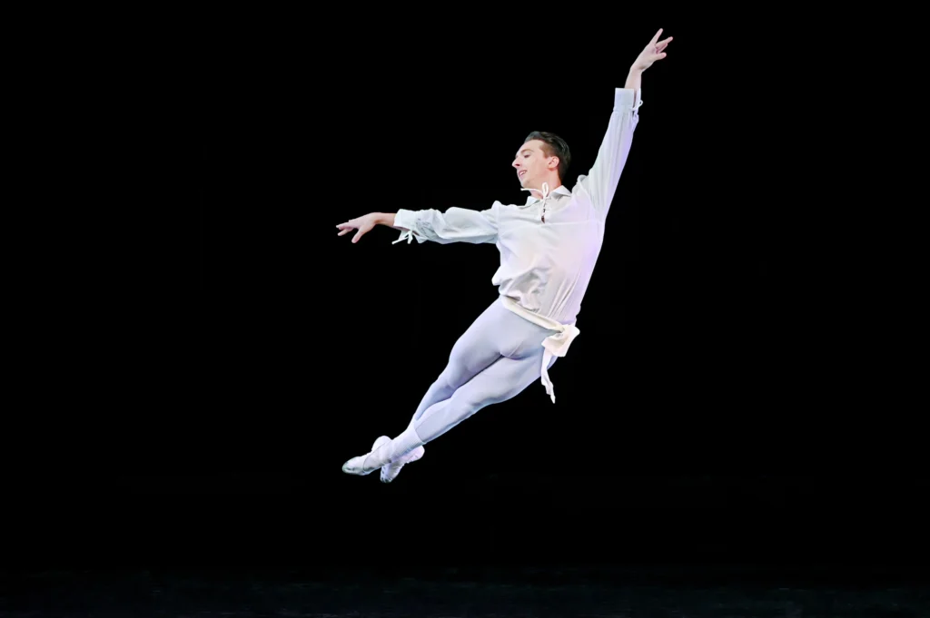 Robert Fulton performs a cabriole derriere in front of a black backdrop, holding his arms in first arabesque and looking out towards the audience. He wears a white long-sleeved blouse, gray tights, white socks and white ballet slippers.