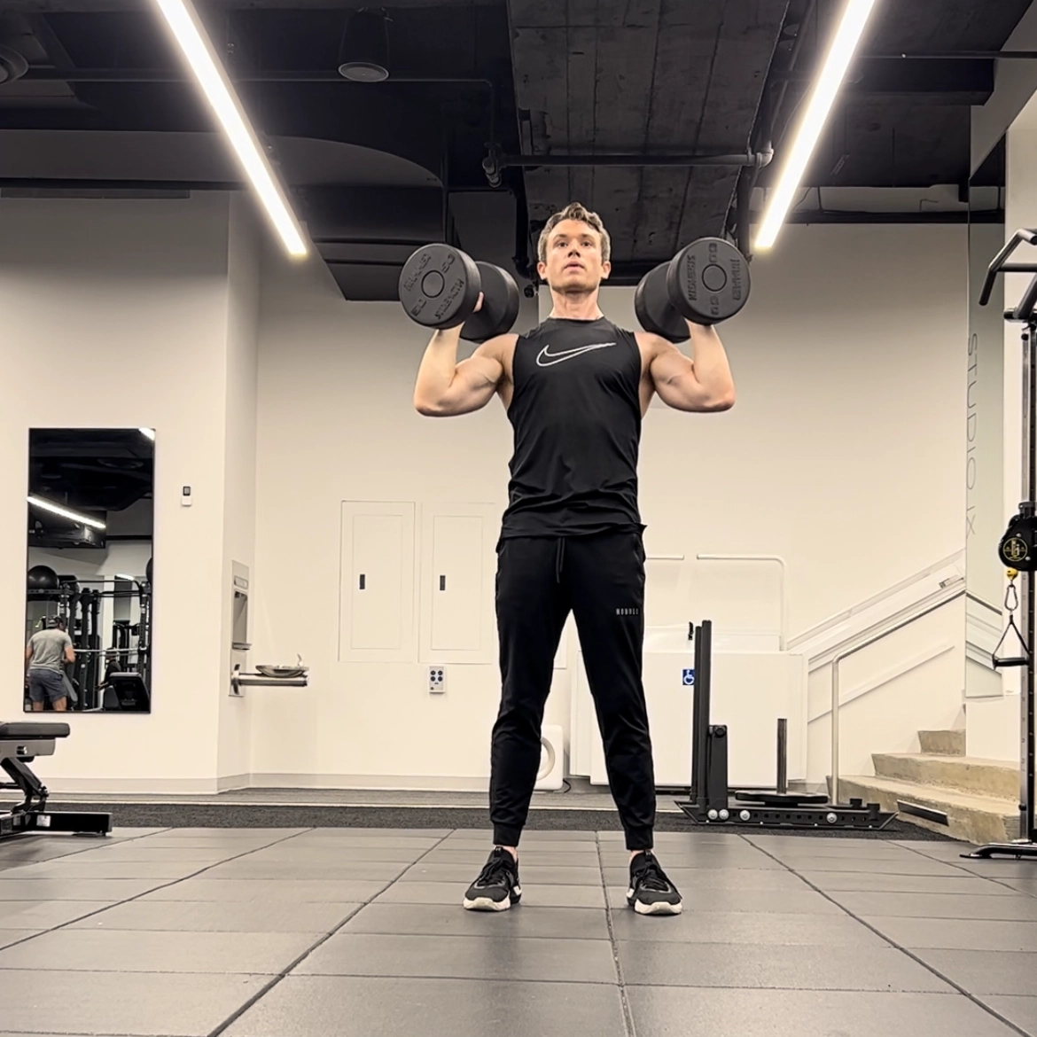 Cameron McCune does a dumbbell exercise at a workout facility. He wears black athletic pants, a black sleeveless shirt and black sneakers. He stands with his feet shoulder-width apart, bends his arms so that his hands are just above his shoulders, and holds a dumbbell in each hand.