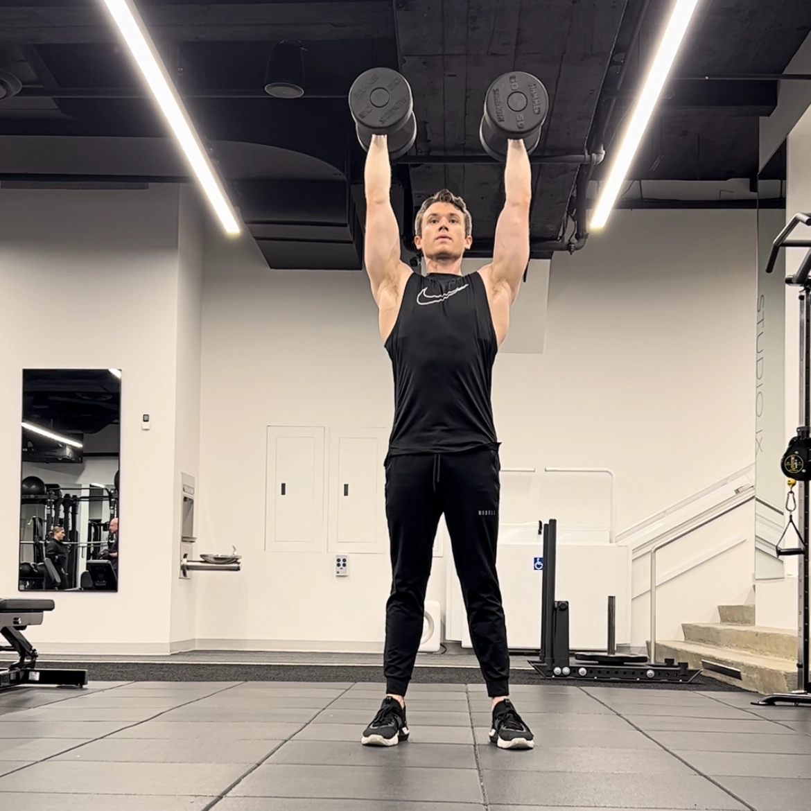 Cameron McCune does a dumbbell exercise at a workout facility. He wears black athletic pants, a black sleeveless shirt and black sneakers. He stands with his feet shoulder-width apart, raises his arms straight above his head, holding a dumbbell in each hand.