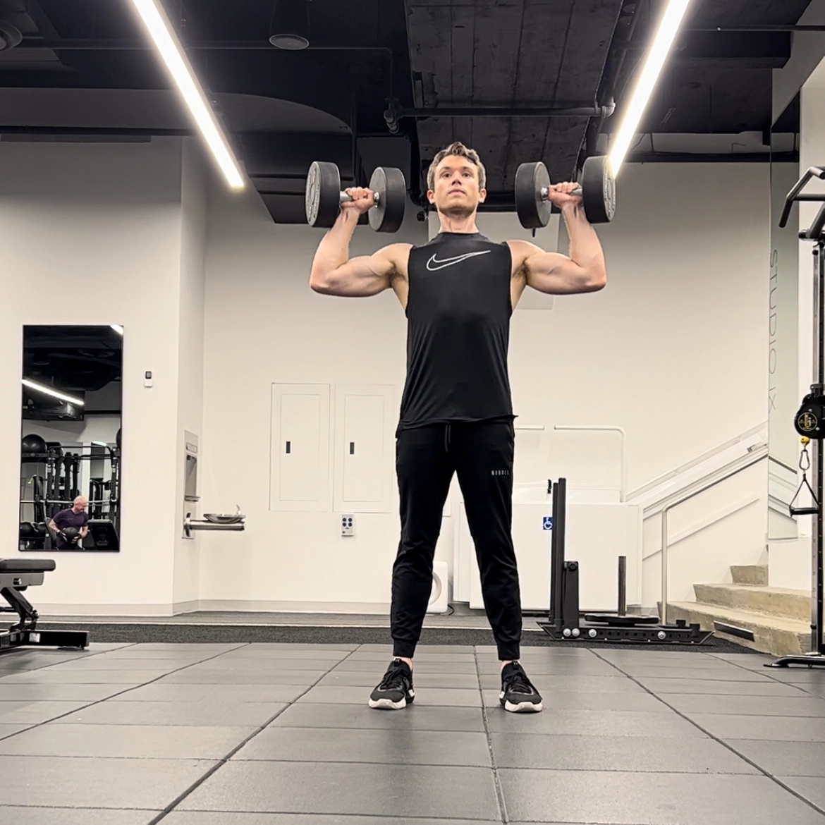 Cameron McCune does a dumbbell exercise at a workout facility. He wears black athletic pants, a black sleeveless shirt and black sneakers. He stands with his feet shoulder-width apart, bends his arms so that his elbows are out to the side and his hands are just above his shoulders, and holds a dumbbell in each hand.