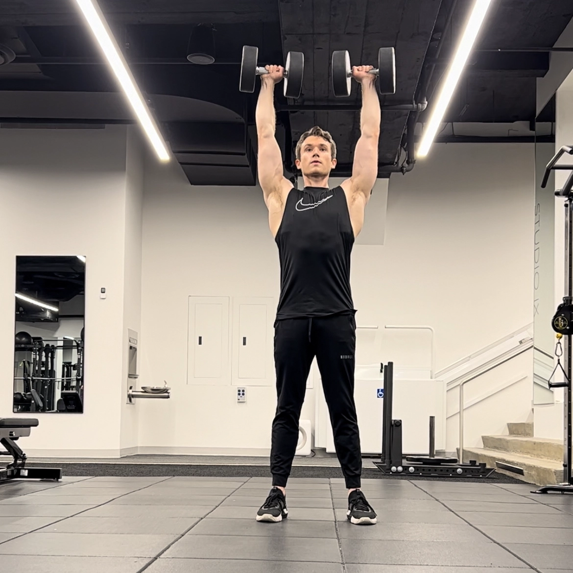 Cameron McCune does a dumbbell exercise at a workout facility. He wears black athletic pants, a black sleeveless shirt and black sneakers. He stands with his feet shoulder-width apart and aises his arms directly above his head, holding a dumbbell in each hand.