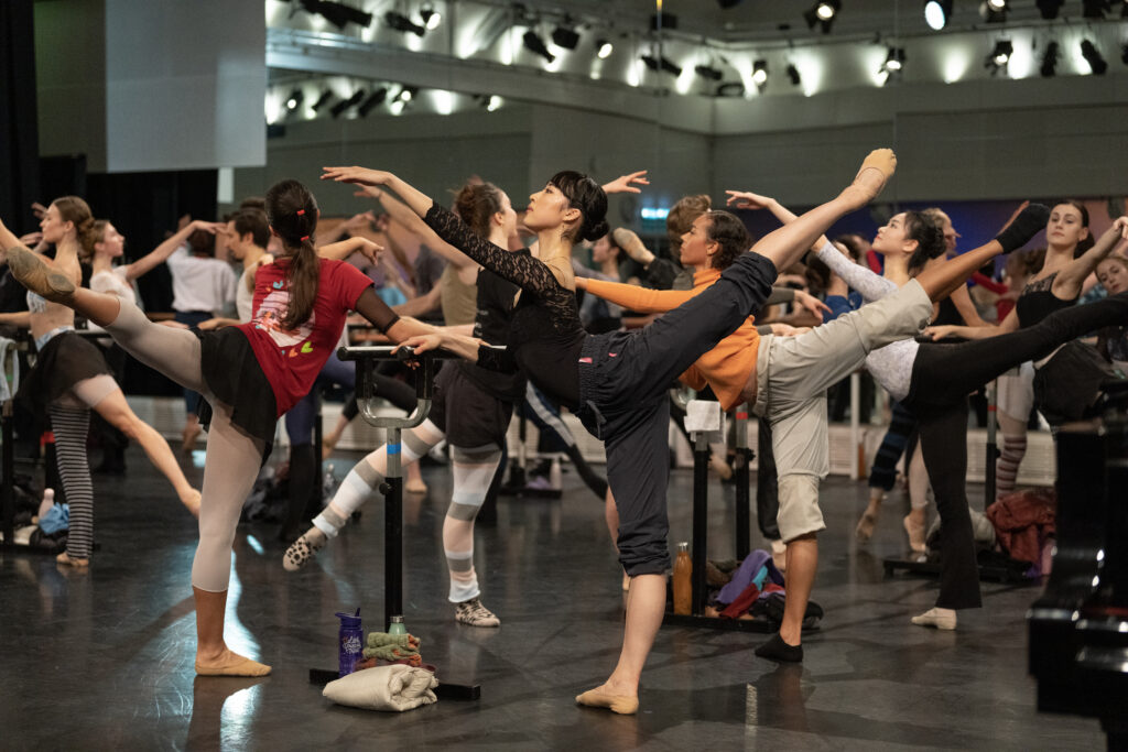Sumina Sasaki in class with The Royal Ballet on World Ballet Day 2022. The dancers all arabesque at the crowded barres in a large dance studio.