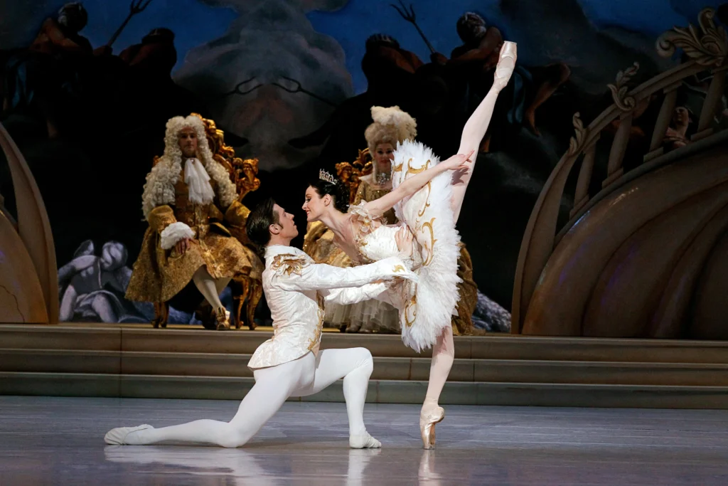 A male and a female dancer perform The Sleeping Beauty's grand wedding pas de deux onstage. She does a penche in toward him, and he lunges forward to support her waist.