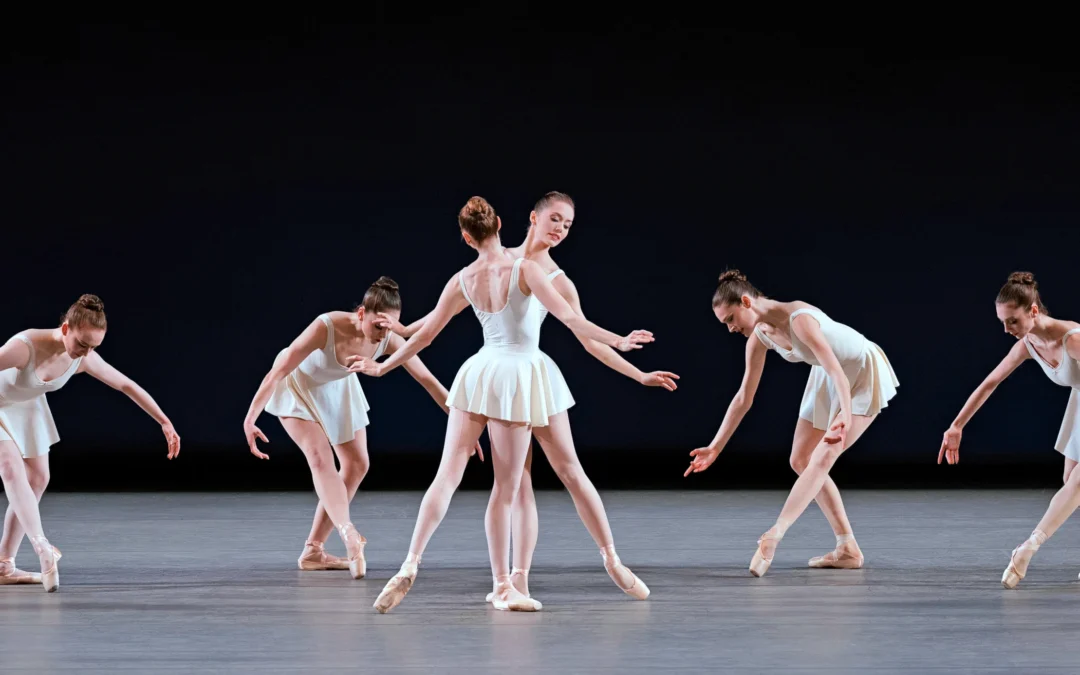 Unity Phelan and Ashley Laracey perform the soloist roles in "Concerto Barocco" onstage. A female corps de ballet dances in a line behind them. All the dancers wear short white ballet dresses and pointe shoes.