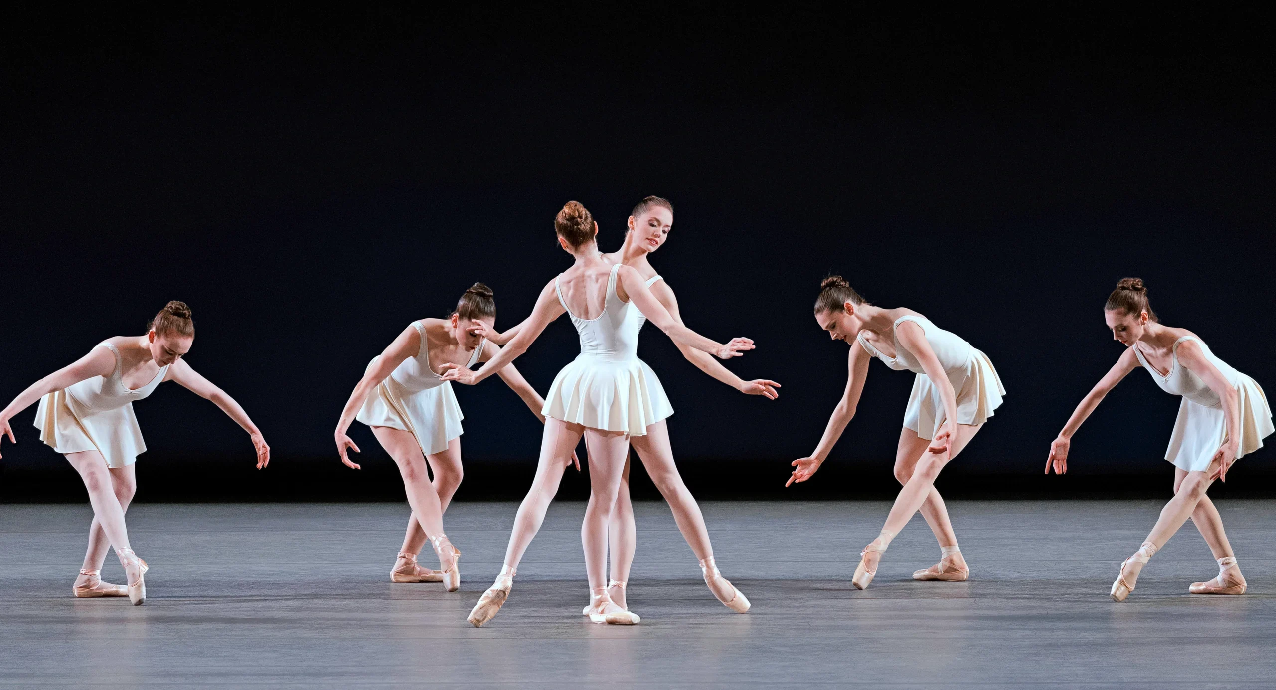 Unity Phelan and Ashley Laracey perform the soloist roles in "Concerto Barocco" onstage. A female corps de ballet dances in a line behind them. All the dancers wear short white ballet dresses and pointe shoes.