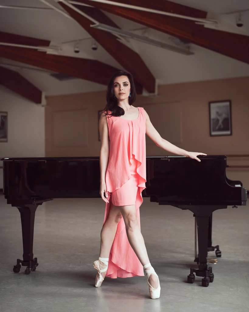Shannon Rugani wears a high-low cut pink dress, standing on pointe in front of a black piano in a large room.