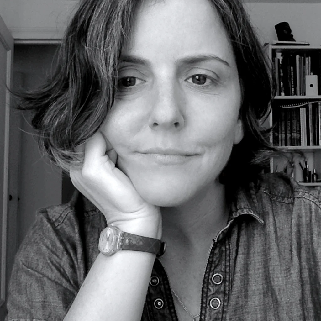 In this black and white photo, Marina Harss is shonw in close-up, leaning her head into her right hand and looking slightly to her right with a small, closed-mouth smile. She wears a denim shirt and watch. IN the background to the left is a tall white bookshelf full of books.