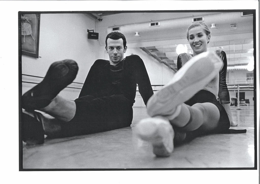 In this black and white photo, Alexei Ratmansky and Gudrun Bojesen sit on a dance studio floor next to each other with their feet outstretched. Each cross their left flexed left foot over their right and smile. Ratmansky wears a dark long-sleeved shirt and dark sweatpants pulled up around his calves, and black ballet slippers. Bojesen wears a long-seleeved leotard, short black ballet skirt, colored tights rolled up and the calf and pointe shoes.