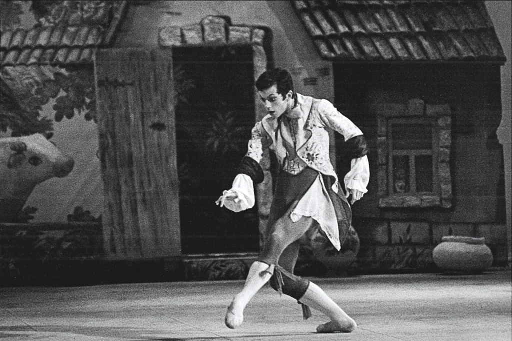 In this black and white photo, Alexei Ratmansky performs onstage in front of a village set. He is taking a step forward, stretching out his left foot and reaching out with his right hand, lookin intently and with surprise at something on the floor. He wears an elaborate 18th century-style jacket with billowing sleeves and tails, breeches, white tights and ballet slippers.