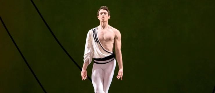 During a performance, Cameron McCune stands onstage with his pointed right foot crossed over his left and his arms held down, palms open towards the audience. He wears white tights and ballet slippers and a white toga-like shirt with one sleeve. McCune looks straight ahead towards the audience.