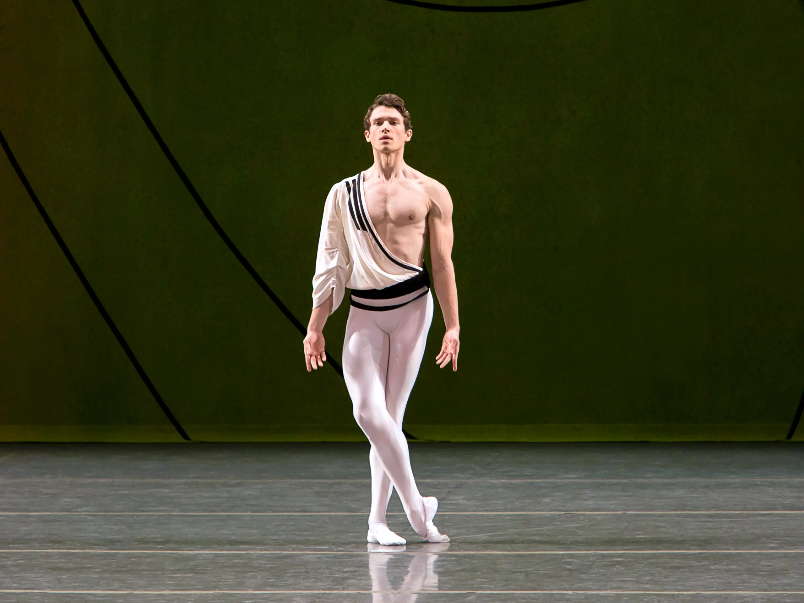 During a performance, Cameron McCune stands onstage with his pointed right foot crossed over his left and his arms held down, palms open towards the audience. He wears white tights and ballet slippers and a white toga-like shirt with one sleeve. McCune looks straight ahead towards the audience.