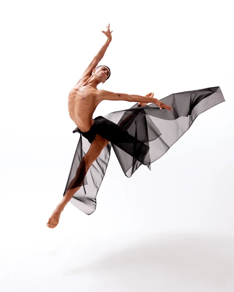 Alberto Andrade wears a long, black tulle skirt that billows behind him as he flies through the air in a contemporary sauté position, his back (right) leg in attitude derriere.