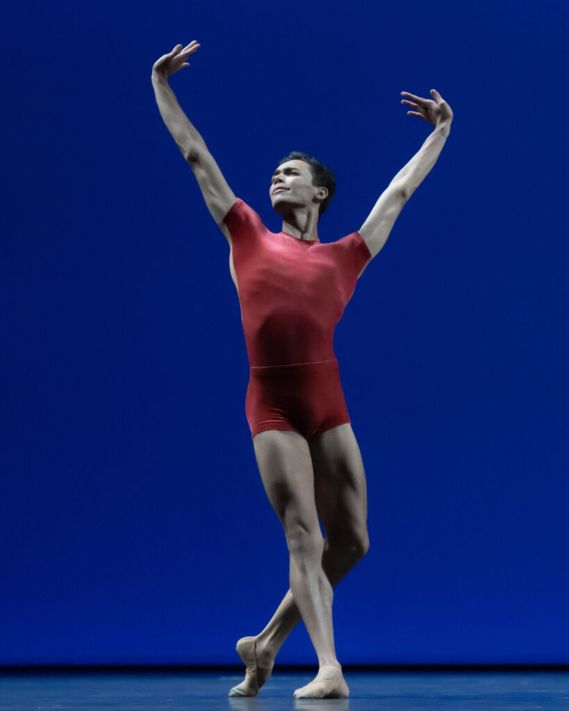 Victor Caixeta, wearing a red biketard, poses onstage in B plus during a performance, his arms lifted into a wide V-shape. He looks proudly out and to his right. He dances in front of a royal blue backdrop.