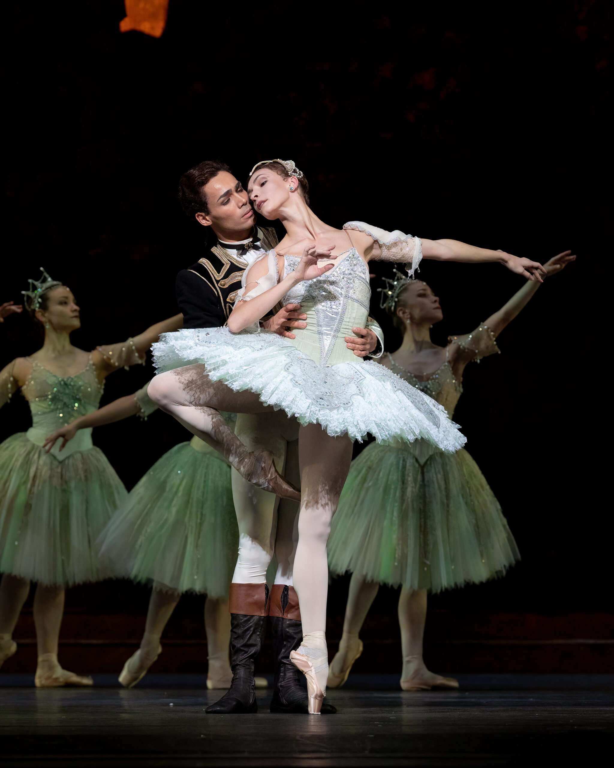 Victor Caiexta and Olga Smirnova dance a pas de deux during a performance of The Sleeping Beauty. Smirnova wears a white tutu and poses on pointe in retiré as Caixeta, wearing a black jacket, white tights and black and bron boots, stands behind her holding her waist. She looks back at him so that their faces are almost touching. Behind them, a trio of corps de ballet dancers in knee-length white tutus embellished with rhinestones and tall crowns pose in tendu derriere effacé.