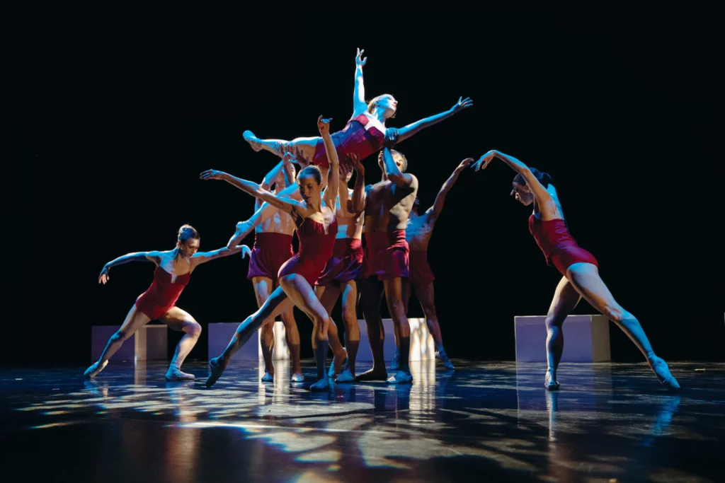 A group if male and female dancers create a tableau onstage during a performance. In the center, three men in red shorts, lift a woman up into the air as she lifts her arms up and out, her legs stretched long behind her. Three women surround them, lunging deeply. All of the women wear red leotards. Behind them upstage are four small, rectangular boxes. The stage is dark, with lights filtered down from above.