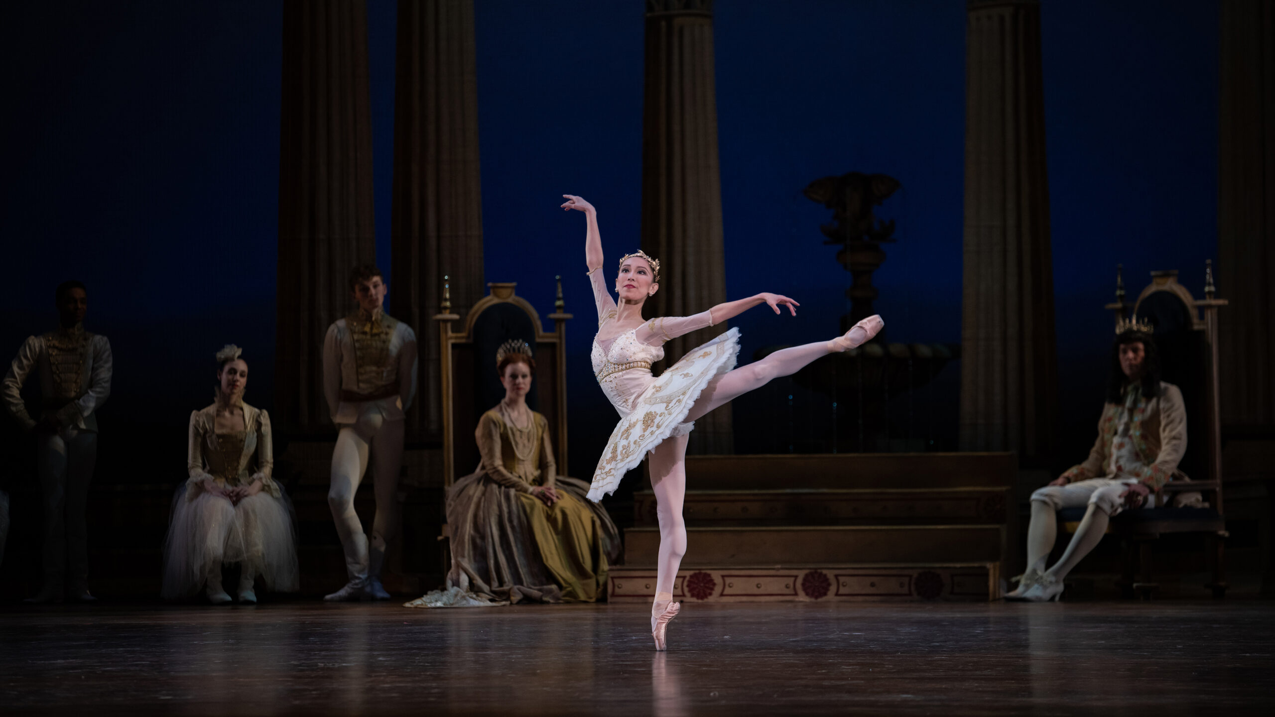 Yuka Iseda does a first arabesque onstage during a performance of Sleeping Beauty, looking out to the audience. She wears a white tutu with gold trim, pink tights and pointe shoes. Behind her, dancers dressed as a medieval royal courtiers sit and watch.