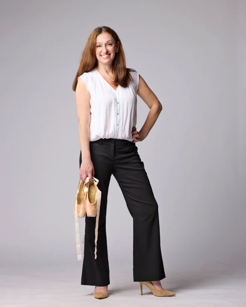 Miriam Landis stands in front of a gray backdrop, holding a pair of pointe shoes in her right hand. She wears black pants, a cap-sleeved white top and tan suede pumps. She smiles for the camera.