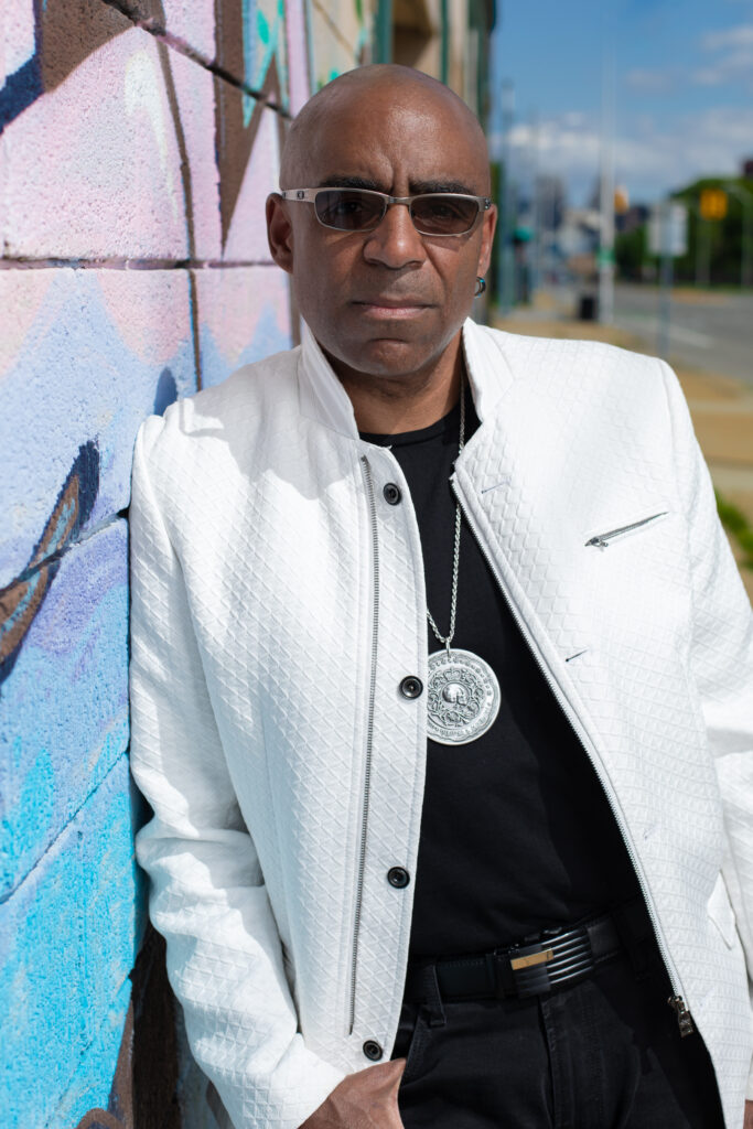 Aaron Dworkin, shown hips-up, leans against a colorfully painted wall outside. He wears dark sunglasses, a black t-shirt, a white jacket, black jeans, and a silver pendant.