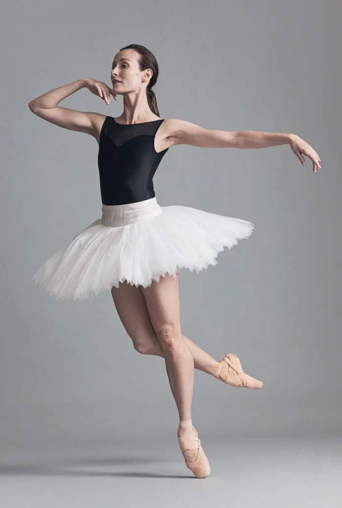 Amber Scott falls forward gracefully in a stylized coupe attitiude position on pointe. her right arm bends gently toward her chin, her left floating to the side. She wears a navy leotard and white practice tutu.