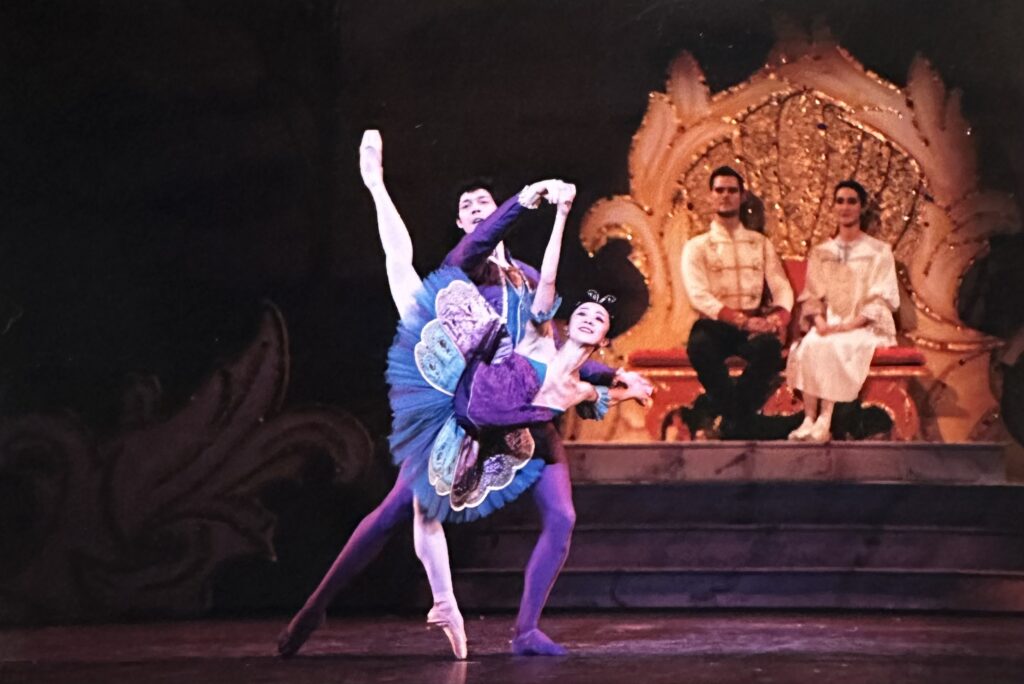 Asuka Sasaki and Christopher Moulton perform a pas de deux together onstage during a performance of The Nutcracker. Sasaki, wearing a purple tutu, does a penché on pointe with her right leg up as Moulton, who wears a purple tunic and tights, poses behind her and holds both of her hands. Behind them, a man and woman sit on a throne and watch.