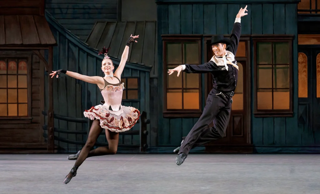 Gilbert Bolden III with Olivia MacKinnon fly through the air in George Balanchine’s Western Symphony. They are jumping in a low cou de pied derriere position, their arms in an extended fourth. They wear stylized western ballet costumes.