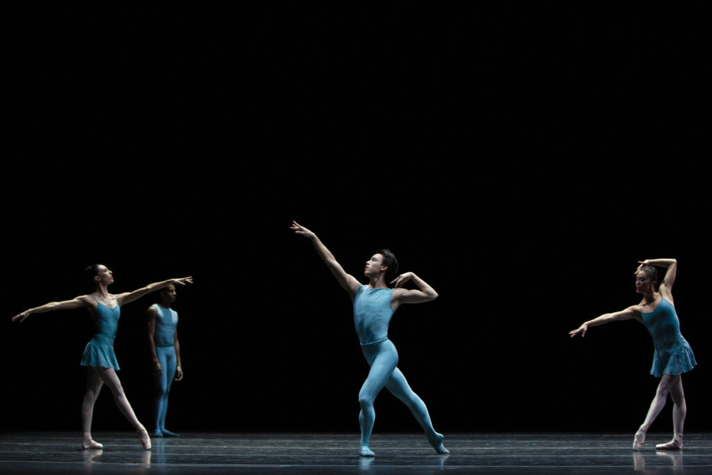 Benji Pearson does a tendu derriere croisé with his right leg back during a performance onstage. He raises hi right arm up and folds his left arm in so that his left hand touches his left shoulder. Pearson wears an all-blue unitard and blue ballet slippers. Behind him, two female dancers in blue pose in tendu croise devant. They perform in front of a black backdrop.