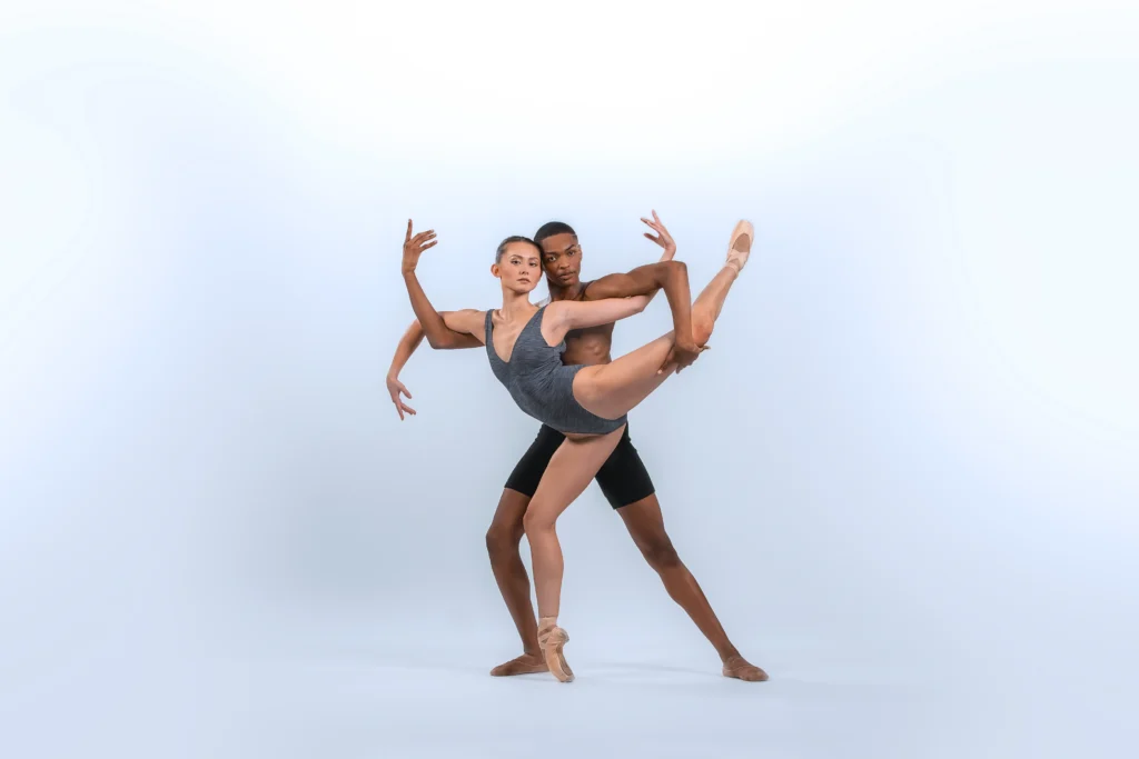 Alessandra Ferrari-Wong poses in an attitude derriere on pointe, bending her right leg in plié and weaving her arms around her partner, LeJeromeny Brown, who stands behind her. Brown holds her left knee in attitude with his left hand and curves his right arm up as she bends her right arm down. He wears black bike shorts, while she wears a gray leotard.