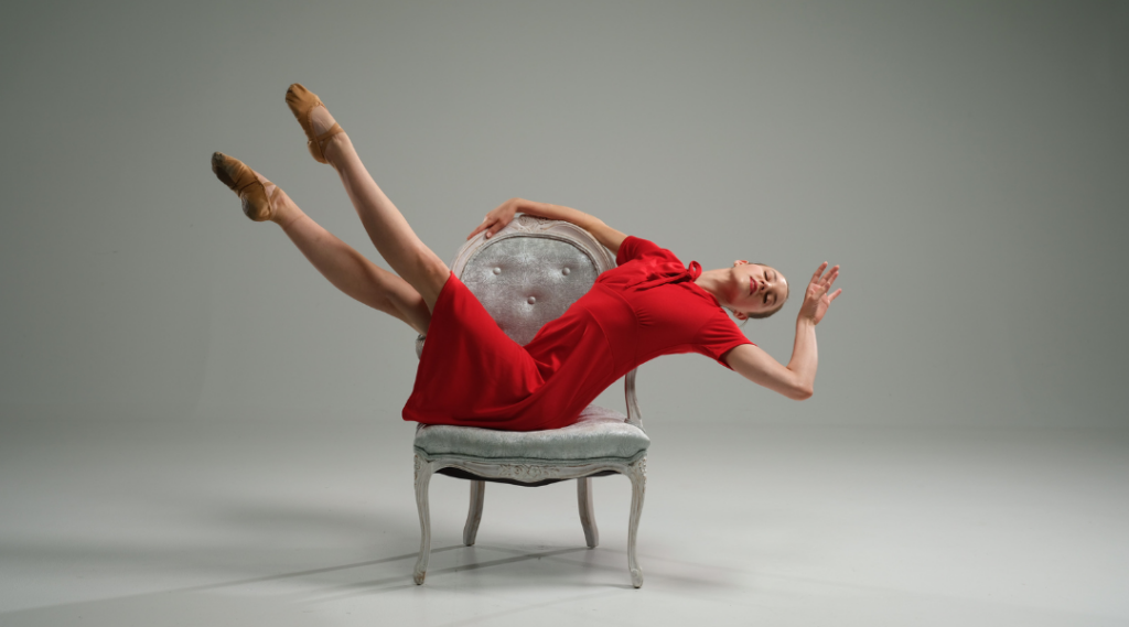 Deos Contemporary Ballet dancer Leah Haggard drapes herself dramatically over a white armchair, wearing a red dress and red pointe shoes. She makes a fake swoon pose with her arm daintily bent and eyes closed.