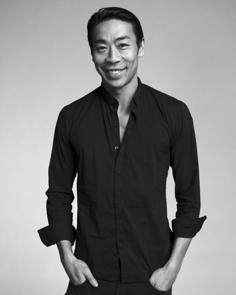 Edwaard Liang poses for a black and white portrait shot, showing him from the hips-up. He smiles wearing a dark button-down, his hands in his pants pockets.