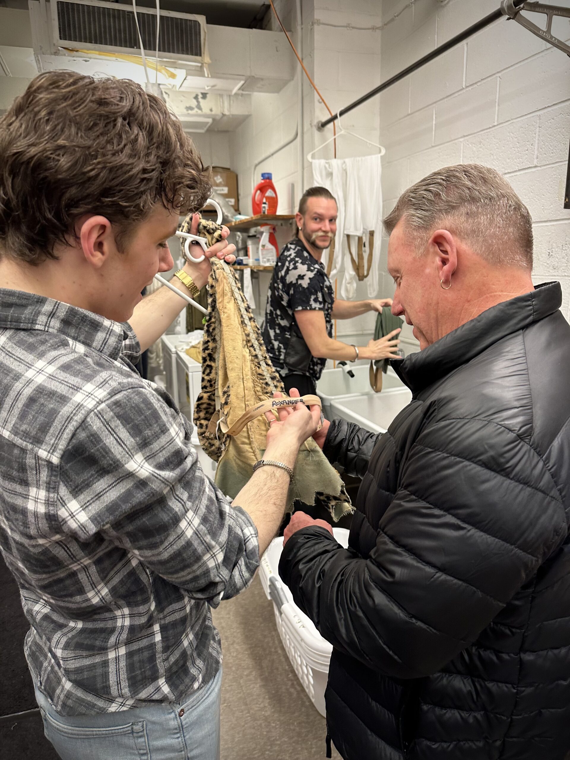 Jake Roxander and David Roxander stand in a theater's costume room. Jake holds up a leopard print costume and holds up a tag that says "Roxander" as his father holds the bottom of the material. They are both wearing street clothing and photographed from behind. A man washing out tights in the sink in the background looks at them and smiles.