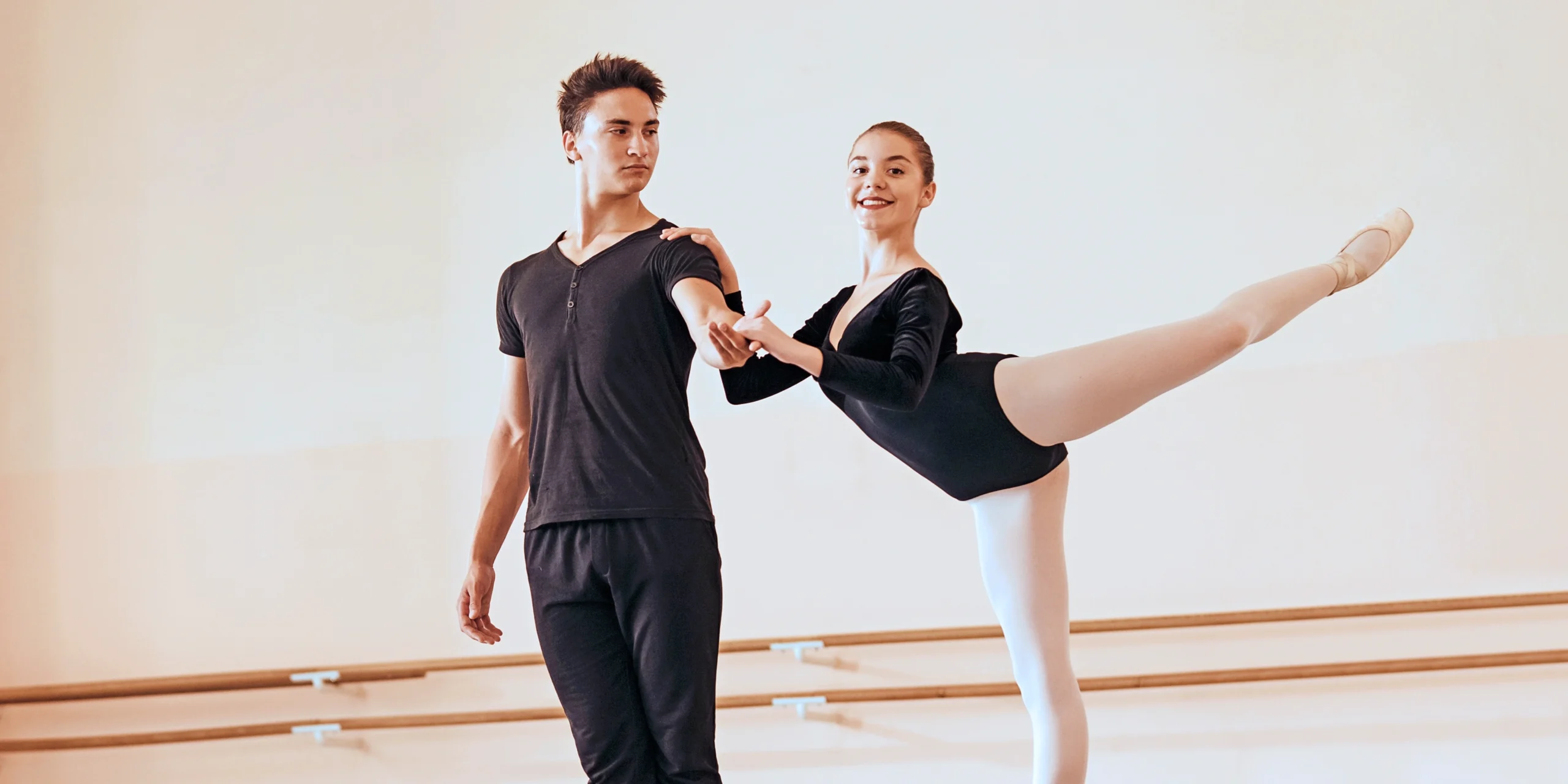 Teenage dancers rehearse a pas de deux in a large dance studio. The girl, wearing a long-sleeved black leotard, pink tights, and pointe shoes, does an attitude derrierre and holds onto the boy's left shoulder and left hand. The boy, in a black t-shirt and black sweatpants, stands to her right and looks at her as she looks out to the camera with a large smile.