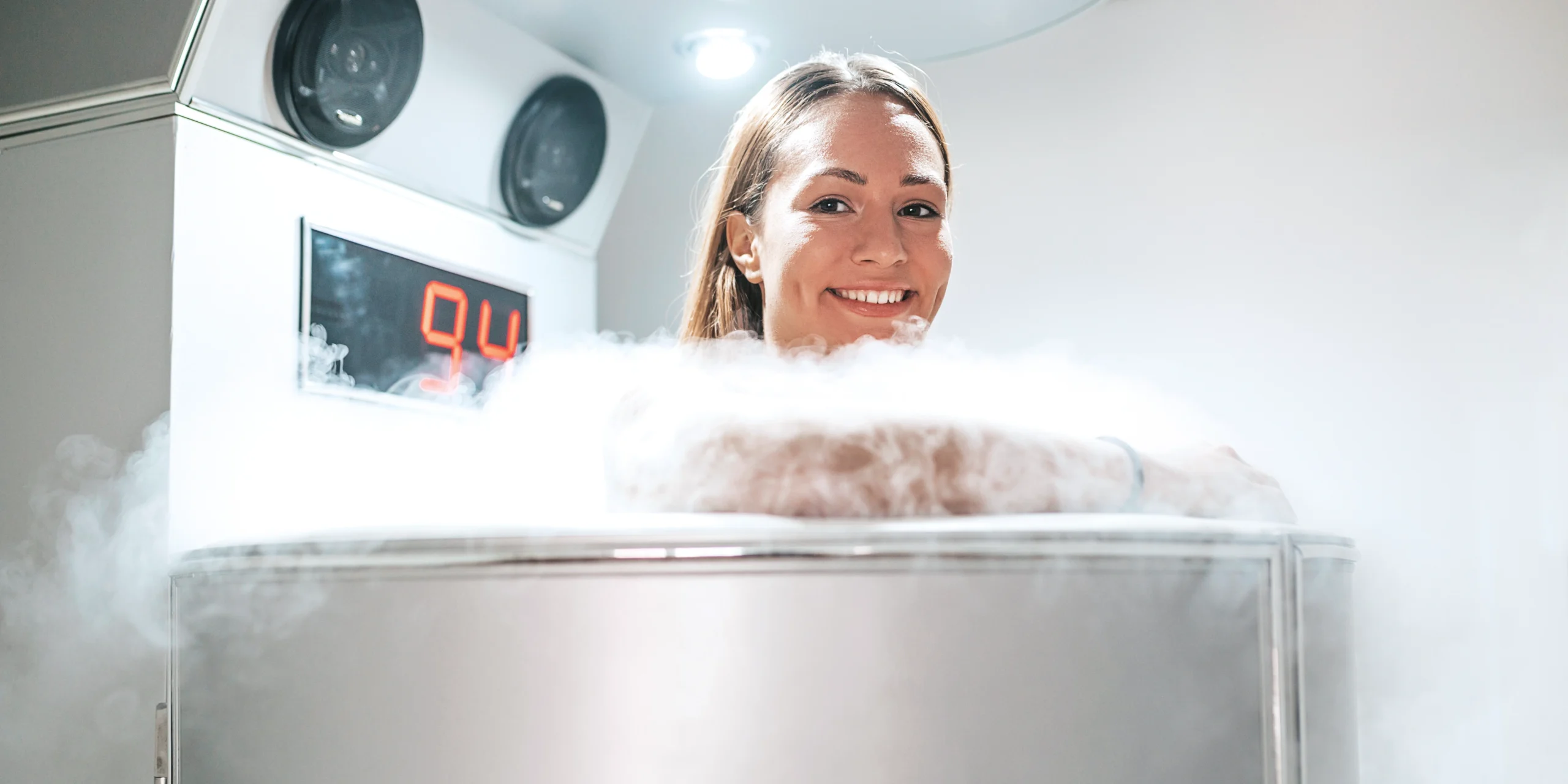 A young blonde woman is shown in a steel cryotherapy chamber. Only her neck and head are visible, and liquid nitrogen swirls around her. She looks towards the camera and smiles.