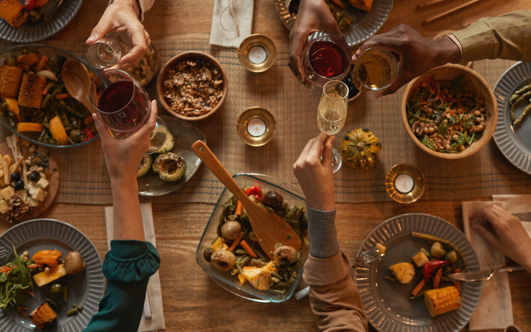 Above view of multi-ethnic group of people enjoying a Thanksgiving feast. They clink glasses and have plates full of food. Dishes of casseroles, potatoes and more are scattered across the tabletop.