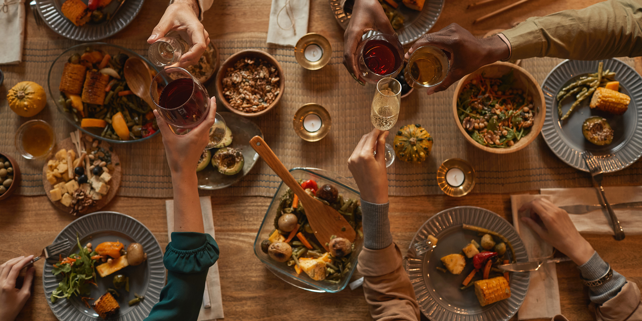 Above view of multi-ethnic group of people enjoying a Thanksgiving feast. They clink glasses and have plates full of food. Dishes of casseroles, potatoes and more are scattered across the tabletop.