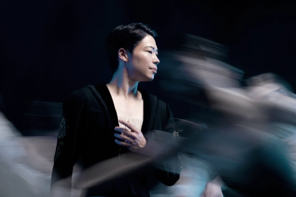 A close-up of Yoshiya Sakura as Albrecht in "Giselle." He poses with his hand over his heart in a black tunic, surrounded by wilis.