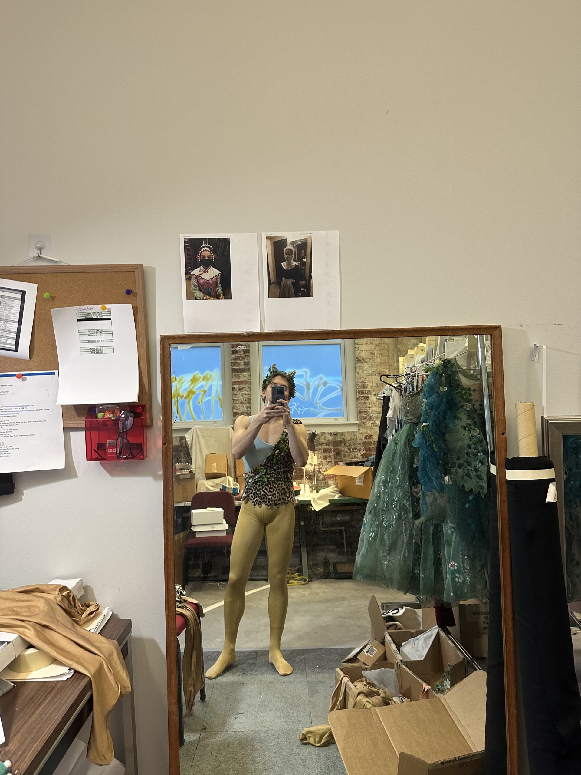 Ashton Roxander takes a selfie in his Puck costume, posing in front of a mirror that's resting on the floor of a wardrobe room. He wears a one-shouldered, leopard-print toga top with leaves trimming the edges, mustard-colored tights, and a headpiece with two small horns. He stands next to a clothing rack lined with blue-green costumes and cardboard boxes along the floor.