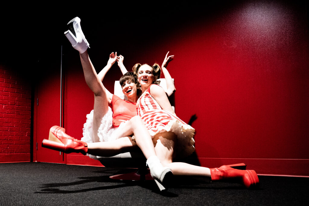 Two female dancers wearing red and white frilly dresses, tights, and high-heel chunky boots pose together in front of a dark red backdrop. They laugh and look at the camera, flinging up their arms and legs.