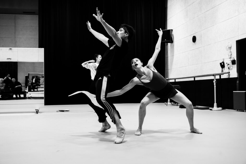 Les Ballets de Monte-Carlo rehearsing <i>L’Enfant et les Sortilèges</i>. In a black and white photo, a female dancer with a surprised face bends sideways in a deep second position plie, holding a leaf-shaped prop underneath the legs of a male dancer, who arches back in surprise.