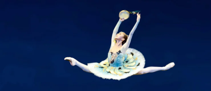 Miko Fogarty does a large saut de chat onstage during a ballet competition. She lifts her arms in high fifth and holds a tambourine in her right hand, and looks up with a triumphant smile. She wears a classical tutu with a blue bodice and blue and white ombre tutu with pastel yellow, green and dark blue trim. She also wears pink tights and pink pointe shoes.