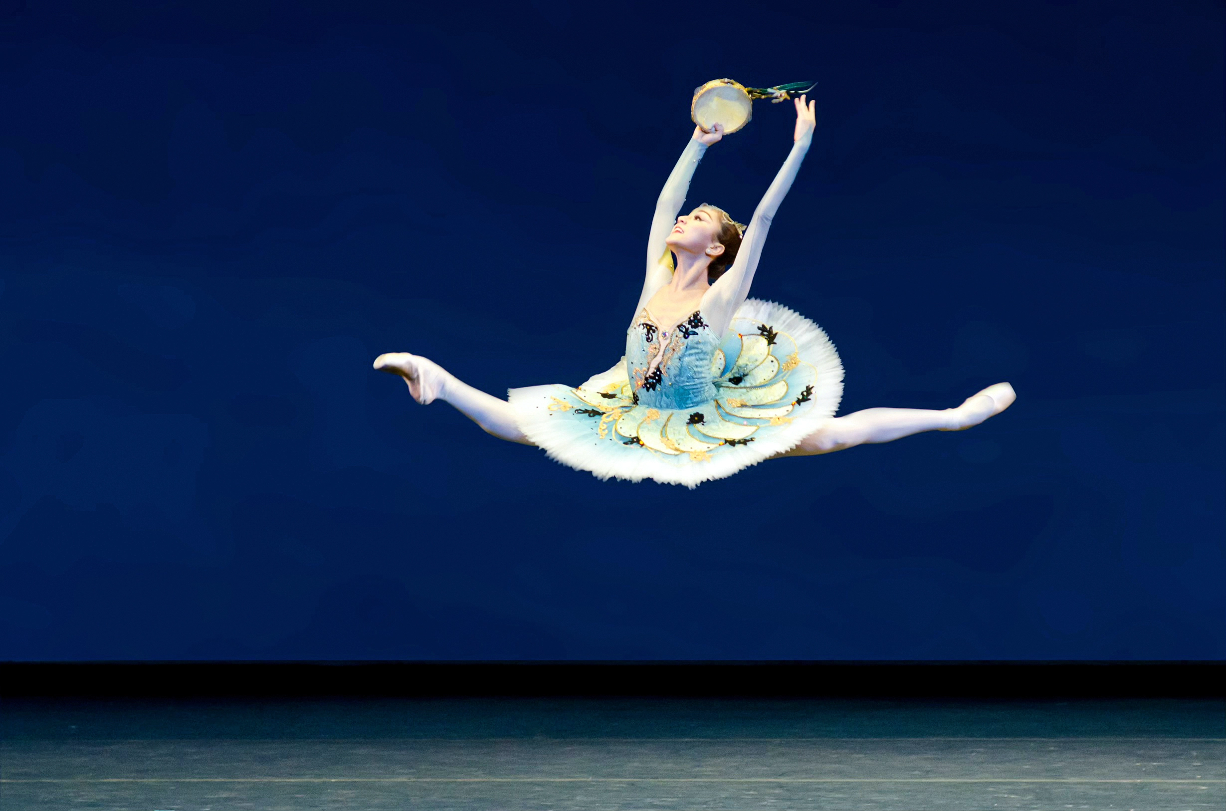 Miko Fogarty does a large saut de chat onstage during a ballet competition. She lifts her arms in high fifth and holds a tambourine in her right hand, and looks up with a triumphant smile. She wears a classical tutu with a blue bodice and blue and white ombre tutu with pastel yellow, green and dark blue trim. She also wears pink tights and pink pointe shoes.