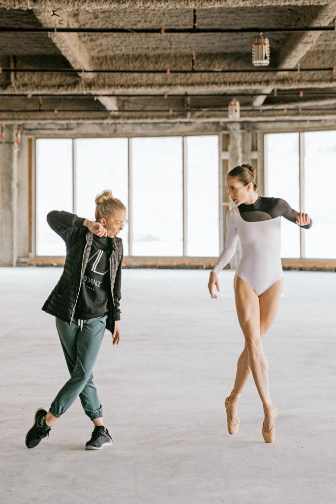 Morgan McEwen and Lauren treat practice dance moves in a large, empty industrial space. Treat, wearing a white leotard with a black left sleeve, stands on pointe, crossing her right foot over her left and looks to McEwen, who is on her right. McEwen, who wears green sweatpants, black sneakers, a dark hoodie and dark t-shirt underneeath, crosses her left foot over her right and looks toward her left.