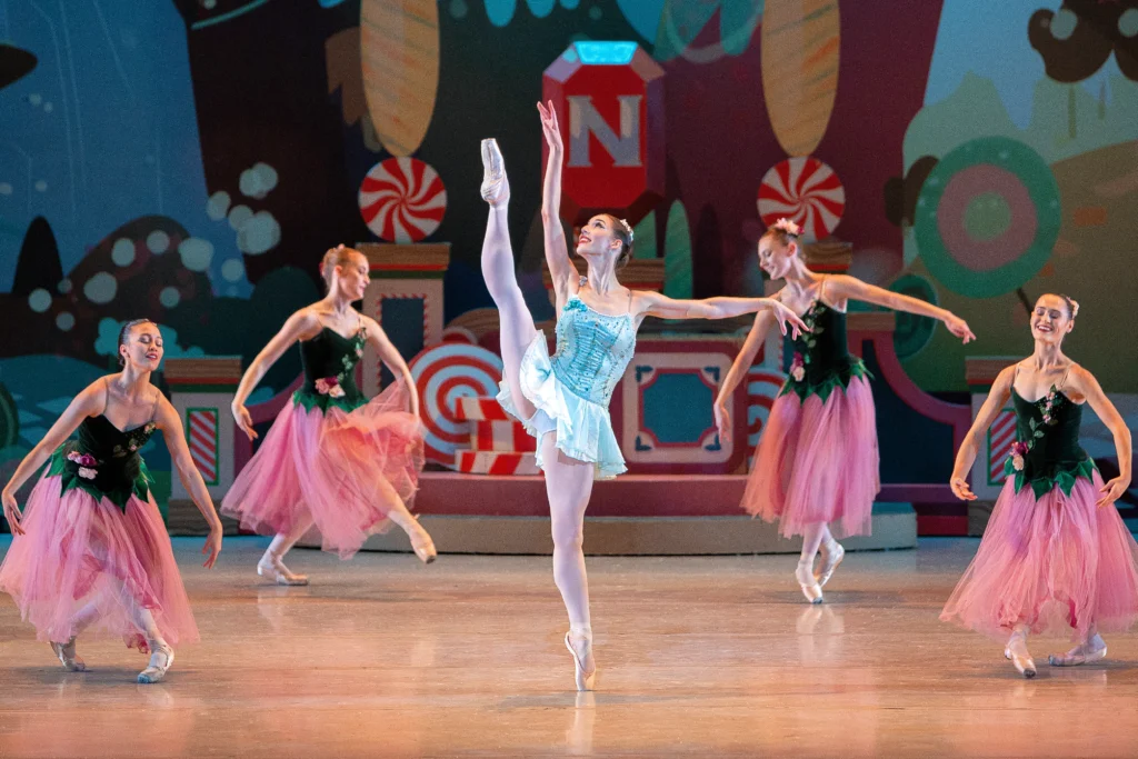 Gabrielle Mengden performs as the Dewdrop Fairy in The Nutcracker ballet onstage. She does a high développé à la seconde with her right leg, lifting her right arm high and her left arm out to the side. She wears a short blue dance dress, pink tights and pink pointe shoes, and is surrounded by four other female dancers in long pink tutus with green velvet bodices.