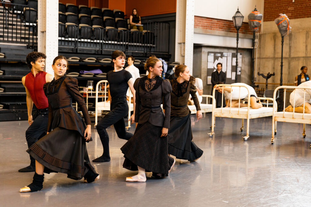 In a large dance studio, five dancers (three women, who stand in front of two men) rehearse a ballet, facing profile towards their right with their legs bent and in parallel. they hold their arms at their sides and look out over their left shoulders. The women wear black Victorian-style dresses and pointe shoes while the men wear rehearsal clothing and ballet slippers. To their left is a row of four twin beds on wheels with a dancer crouched on each one.