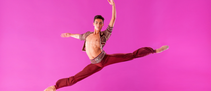 Victor Caiexta does a large sissone in front of a pink backdrop. He looks directly at the camera with a small, closed-lip smile. His legs are split, and he holds his curved left arm up and his right arm out to the side. He is shirtless, wearing a burgundy and silver bolero jacket and burgundy harem pants, and tan ballet slippers.